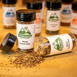 Trailhead brand spice jars on a wooden tabletop, with one jar of black garlic salt tipped over and its contents spilled in the foreground. Other jars are standing upright in the background, and the scene is softly lit with shallow depth of field.